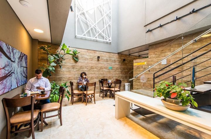 Espaço café da clínica com mesas de madeira, plantas e pé-direito alto